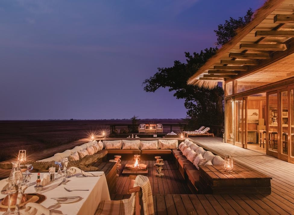 Sunken outdoor lounge with fire pit and dining table on a wooden deck overlooking the Zambian savanna at dusk.