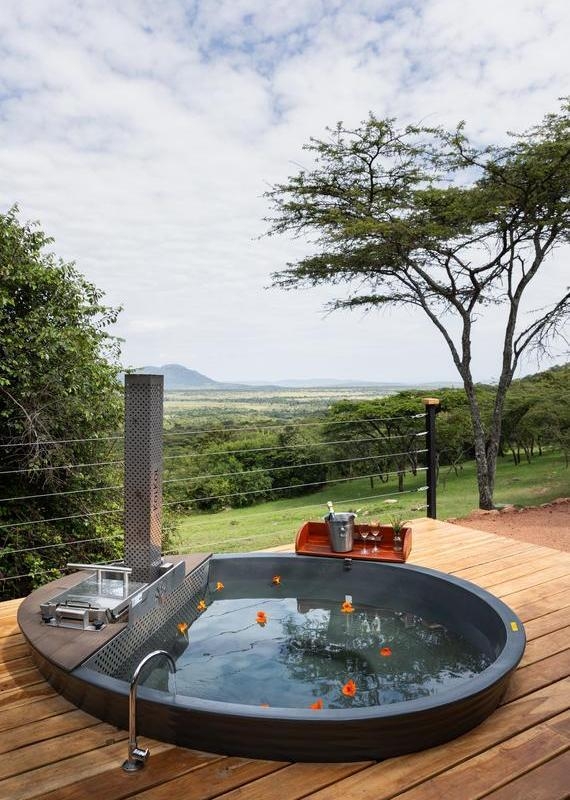 Outdoor soaking tub labeled KolKol on a wooden deck with floating orange flowers, overlooking a vast green savanna.