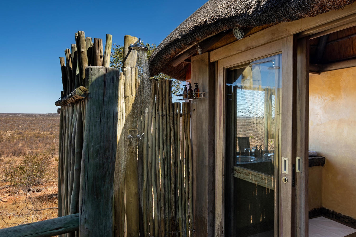 Rustic outdoor shower at Ongava Lodge made of wooden poles overlooking an expansive savanna landscape.