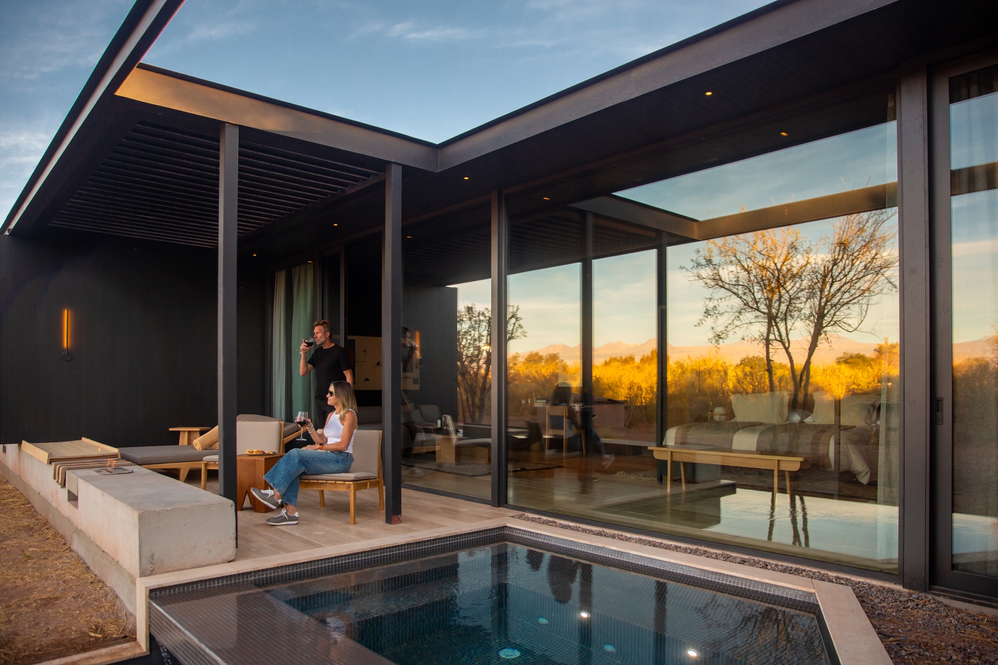 A couple sits on a wooden terrace next to a small pool outside a modern glass-walled building during evening twilight.