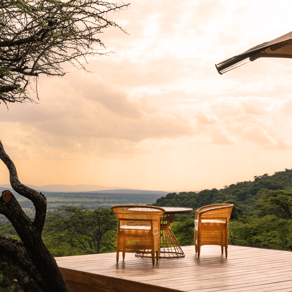 Wooden deck with two wicker chairs and a small table overlooking an expansive green savanna under a cloudy, warm sky.