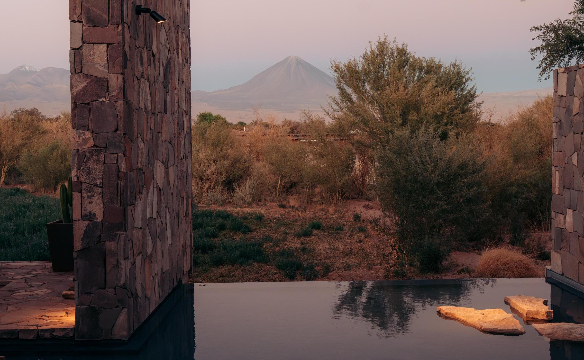 Stone architectural walls and a still pool of water reflecting a hazy pink sunset sky over desert shrubs and a volcano.
