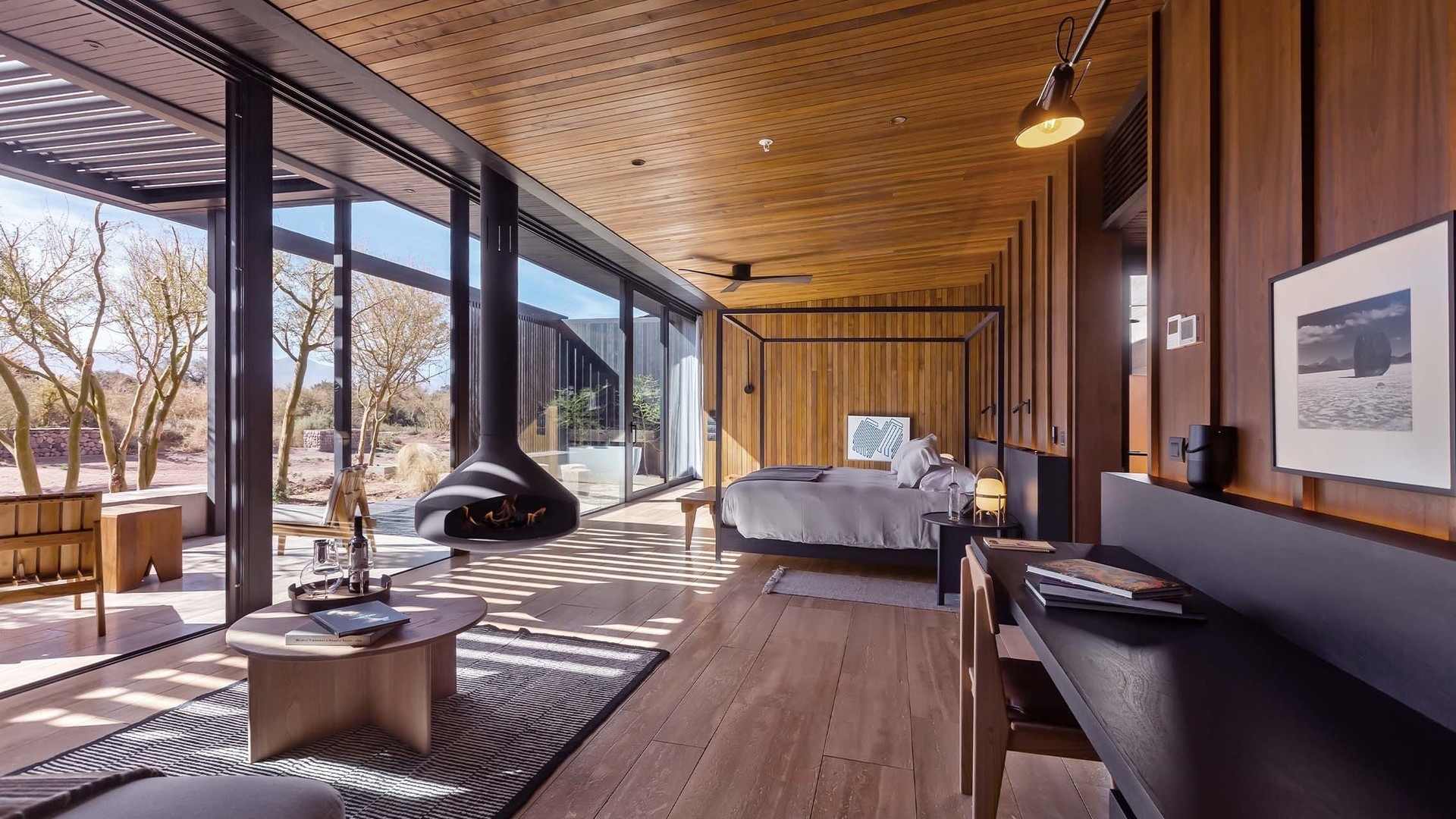 Hotel room with wood paneling, a large bed, modern seating, and a large window looking out at a desert landscape.