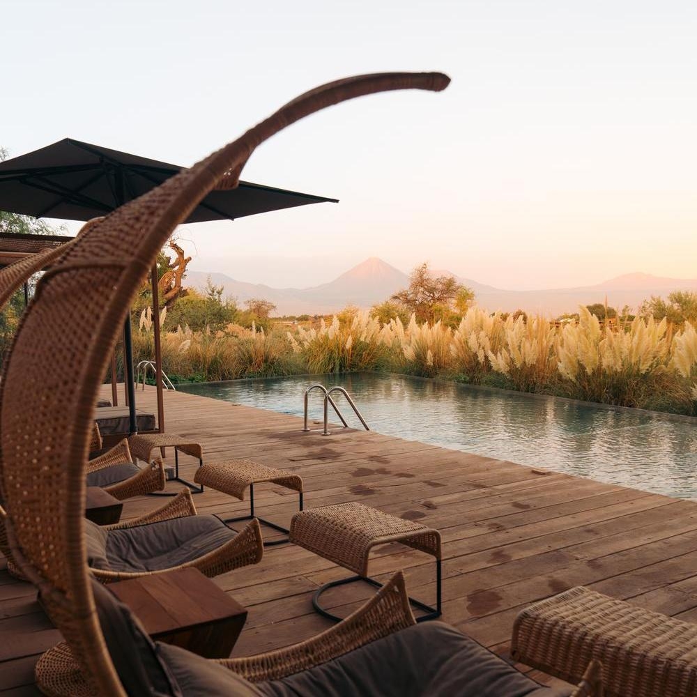 Wooden pool deck with woven lounge chairs facing a swimming pool and a distant volcano peak under a soft sunset sky.