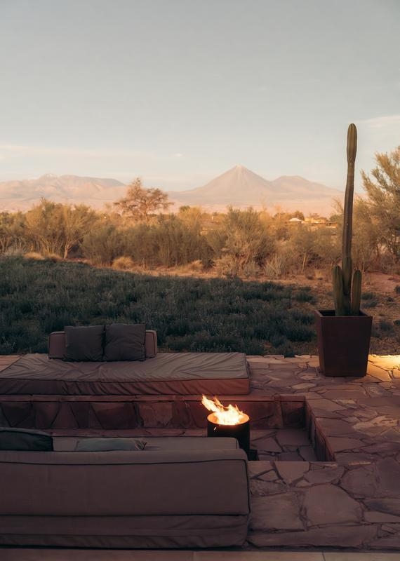 Open-plan hotel suite with wood floors, a hanging fireplace, a bed, and floor-to-ceiling glass doors showing a desert yard.