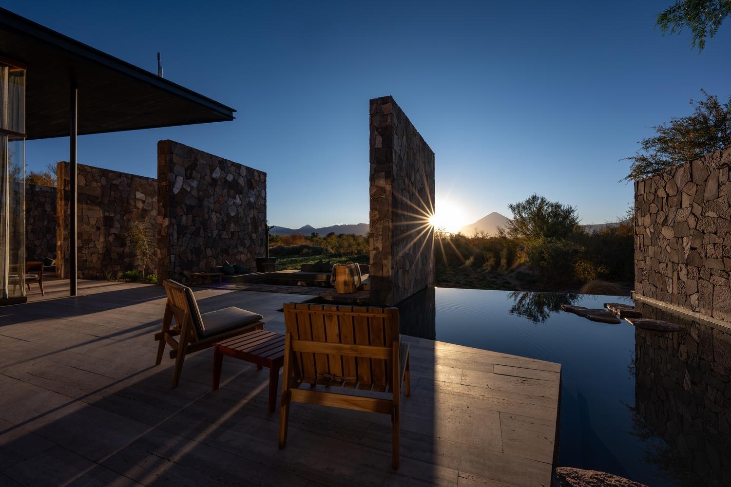 Sunburst appearing past stone privacy walls onto an outdoor patio with wooden chairs next to a calm reflecting pool.