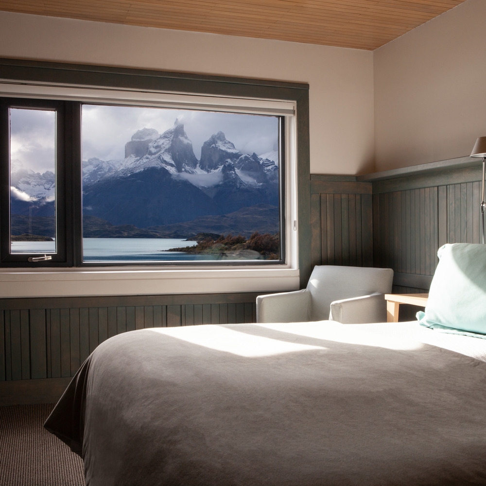 A cozy hotel bedroom at Explora Patagonia with a large window framing dramatic, snow-dusted mountain peaks.