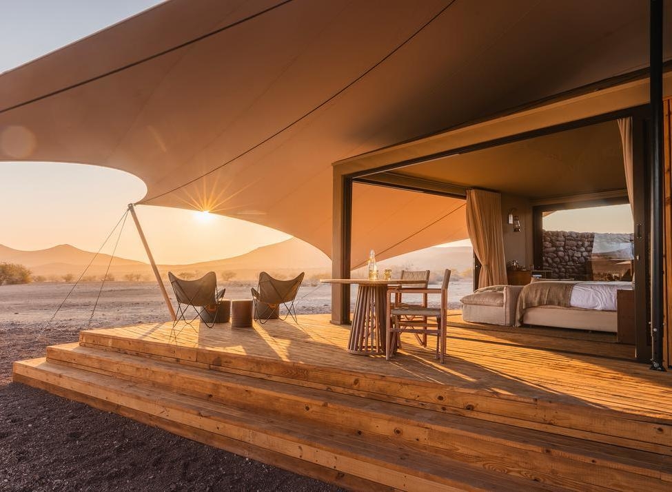 Tented guest suite at Wilderness Desert Rhino Camp with a wooden veranda, lounge chairs, and desert views at dawn.