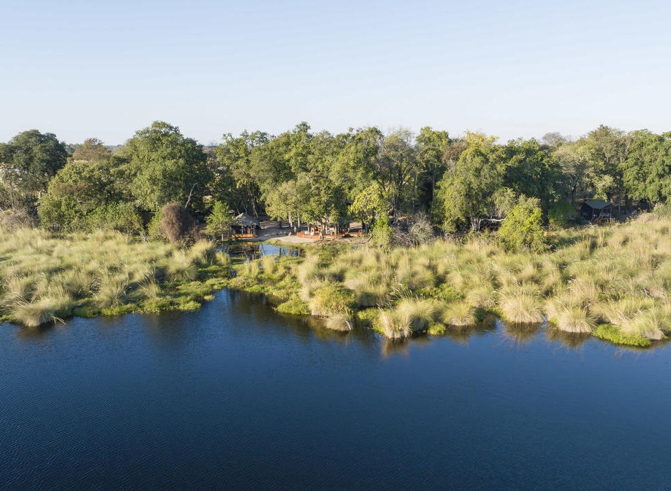 High angle view of a safari camp hidden in trees next to a blue waterway and grassy marshland.