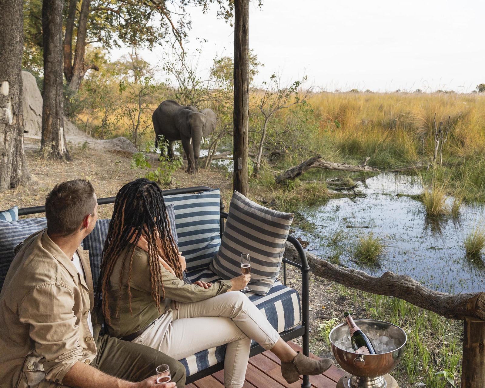 Two people on a safari tent balcony watch an elephant standing near water and tall grass.