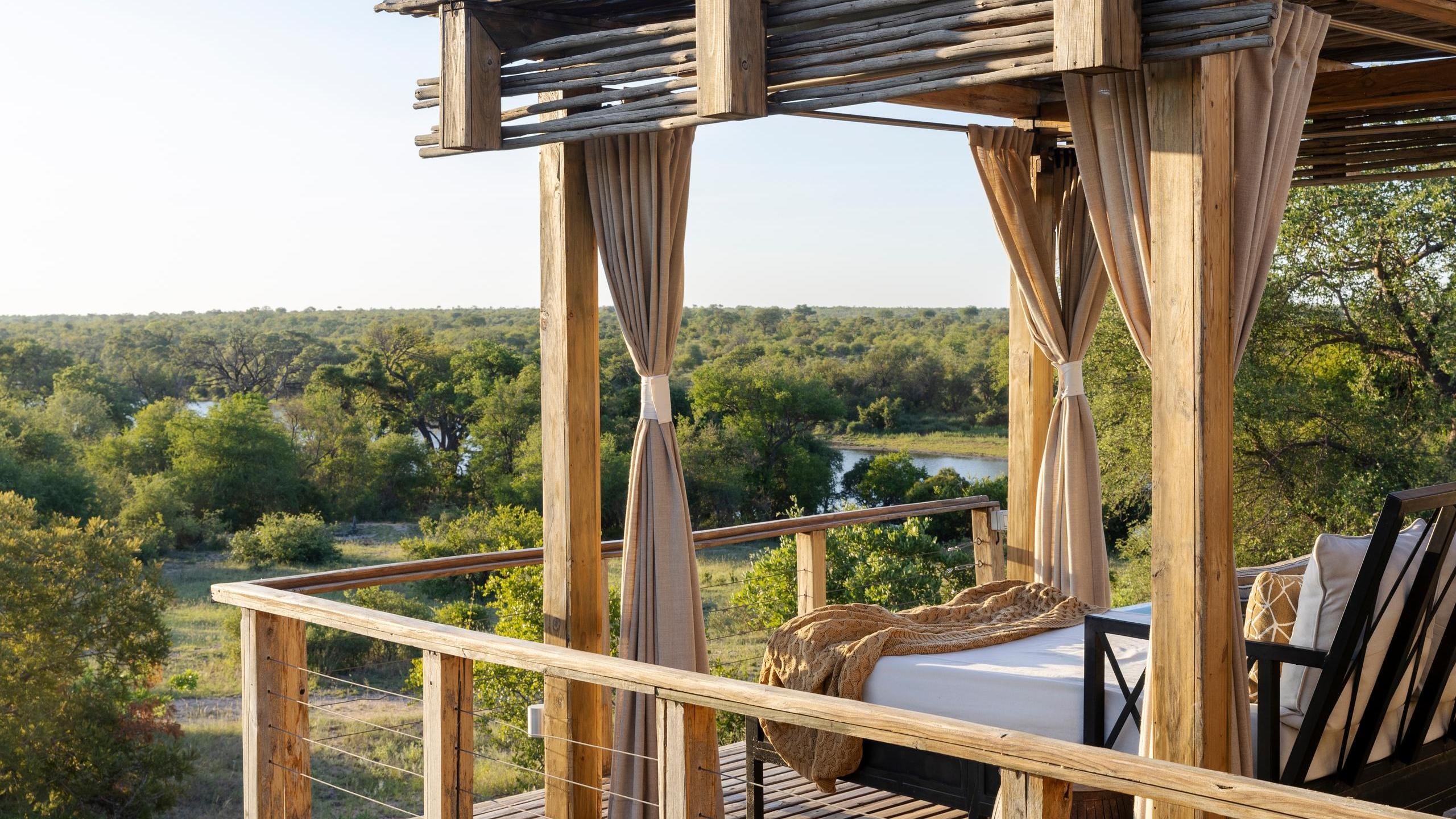 A shaded daybed on a wooden balcony overlooking the river and trees at Simbavati Hilltop Lodge.