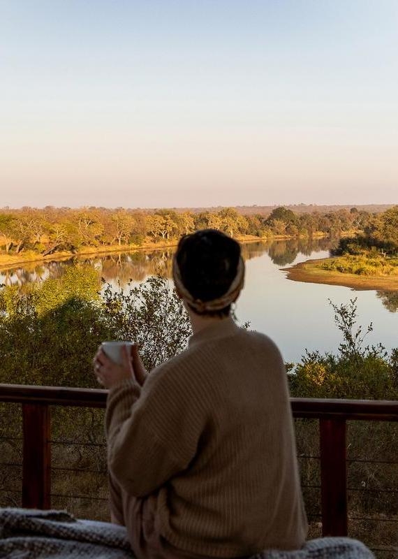 Rear view of a person looking at a calm river from a wooden balcony at Simbavati Hilltop Lodge.