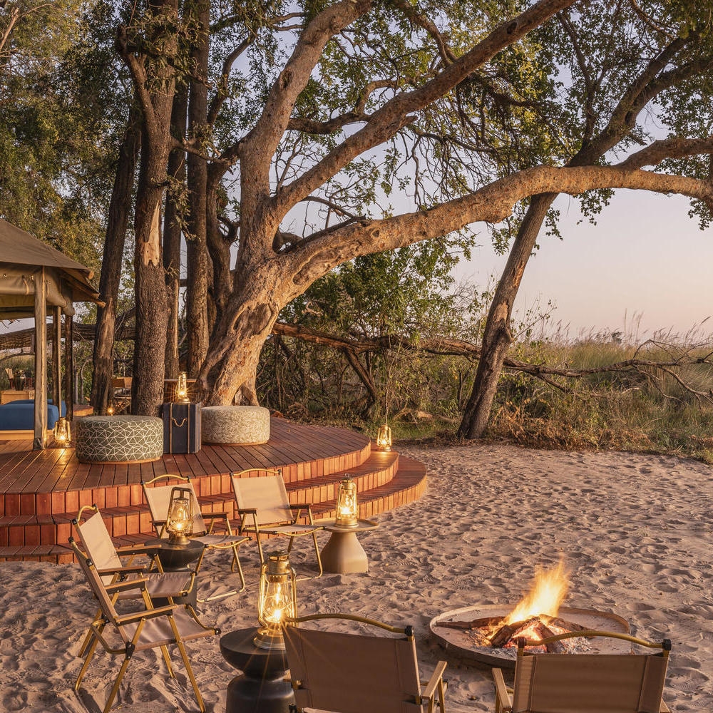 A small campfire surrounded by tan chairs and lit lanterns on a sandy area next to a wooden platform.
