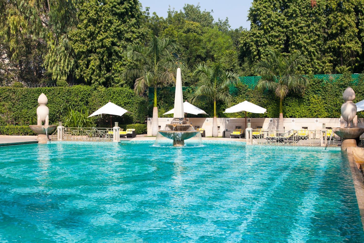 A large blue swimming pool with a tiered stone fountain and lounge chairs under white umbrellas surrounded by greenery.