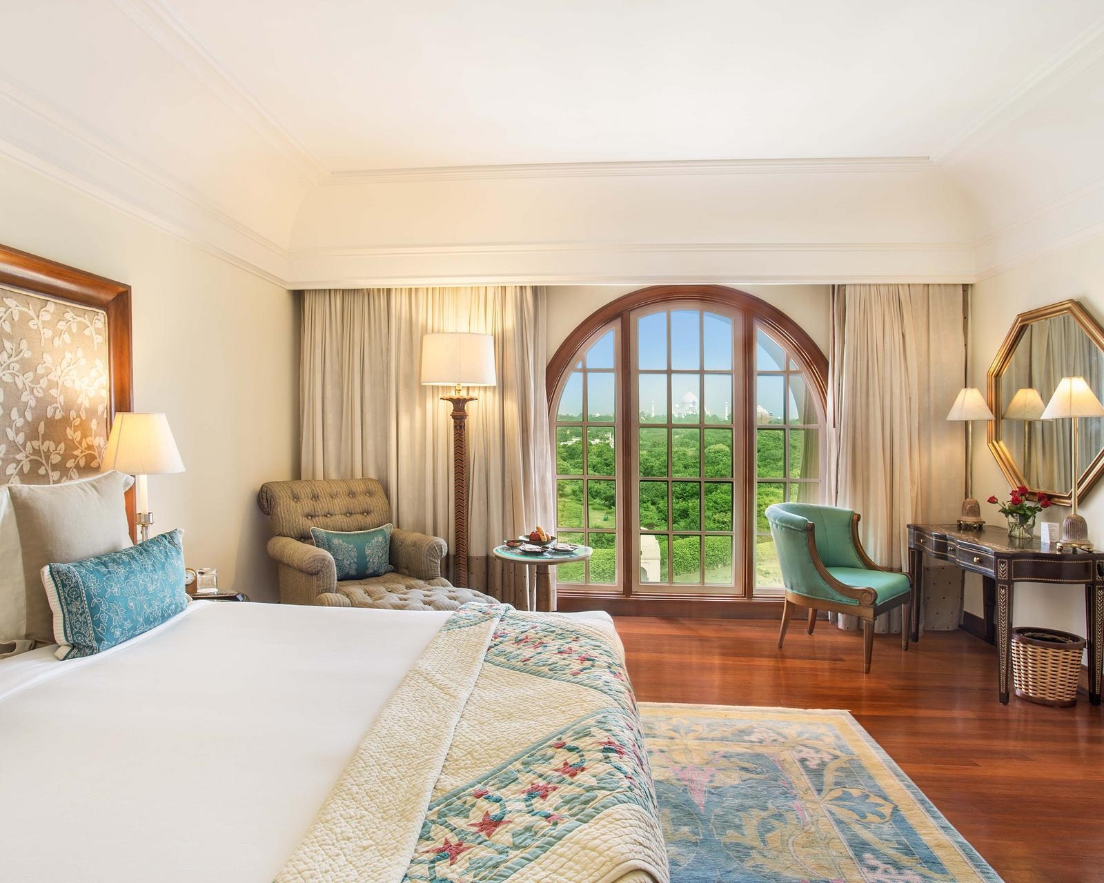 Hotel bedroom with an arched window showing a view of the Taj Mahal over green trees.