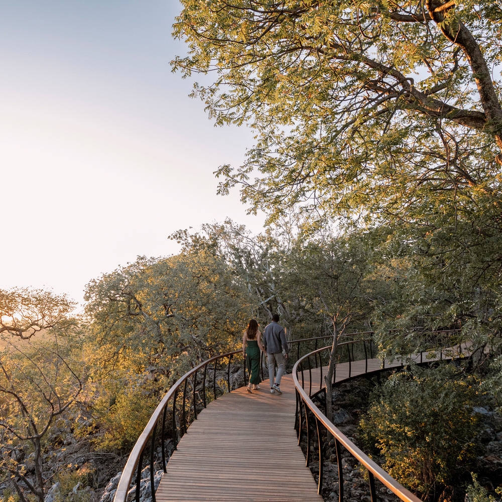 A couple walks hand-in-hand along a winding wooden walkway surrounded by green trees at sunset.