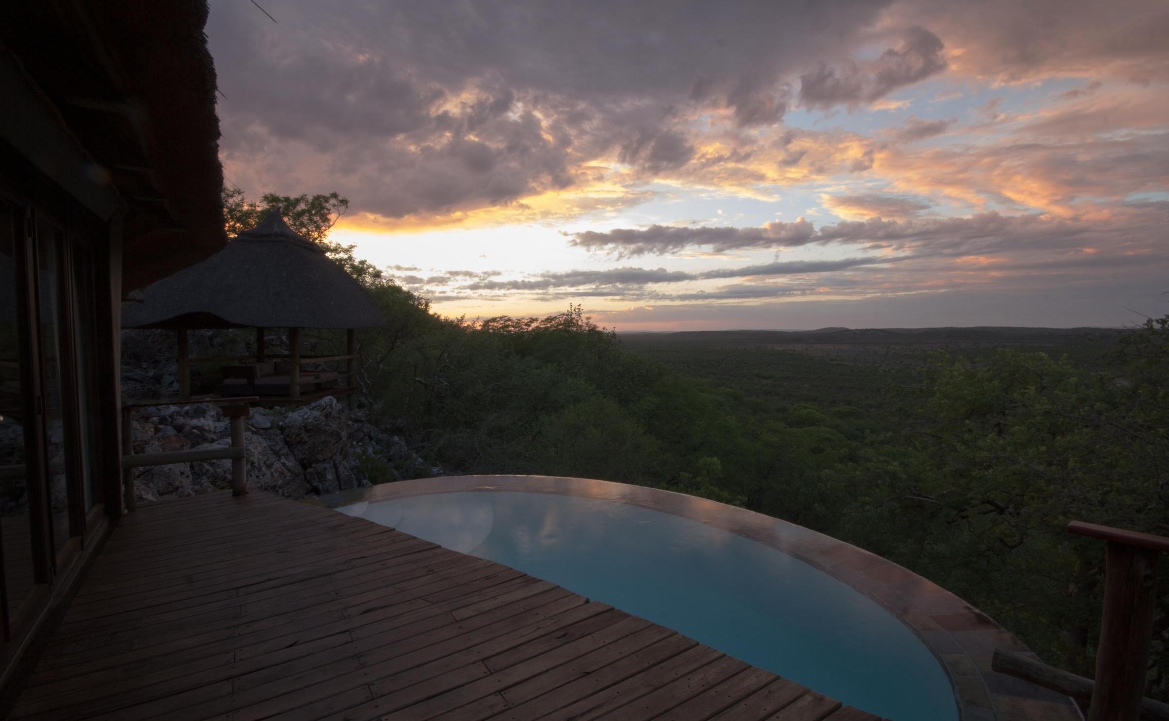 A wooden deck with a curved infinity plunge pool overlooking a vast valley at sunset.