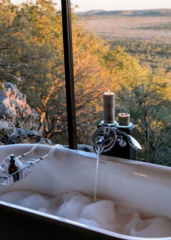 Bubble bath in an outdoor bathtub overlooking a vast wilderness landscape at sunset with lit candles.