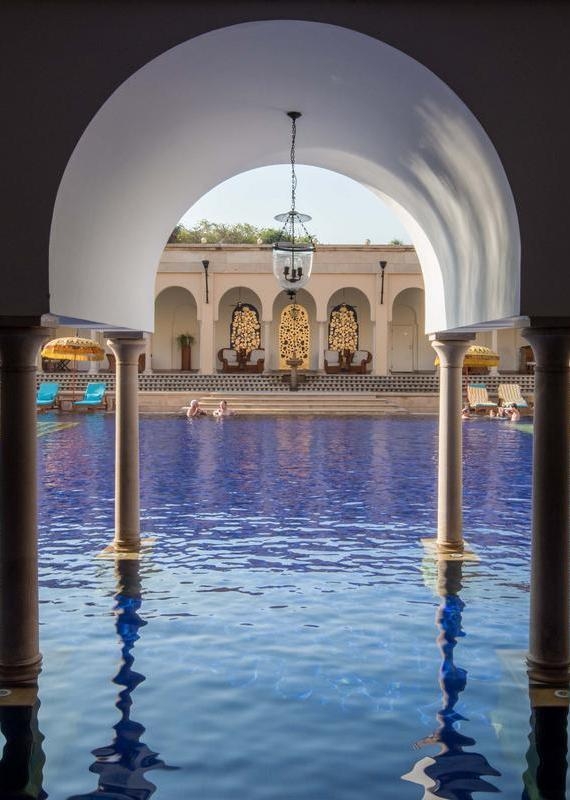 View through an archway of a large blue swimming pool and stone pavilions at The Oberoi Amarvilas.