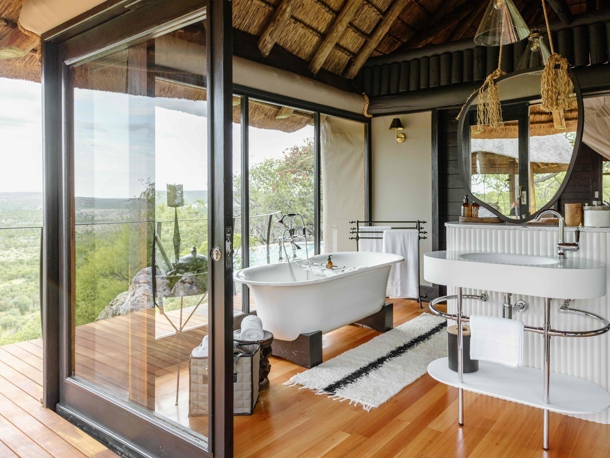 Luxury bathroom with a white freestanding bathtub and vanity next to open glass doors overlooking a wilderness landscape.