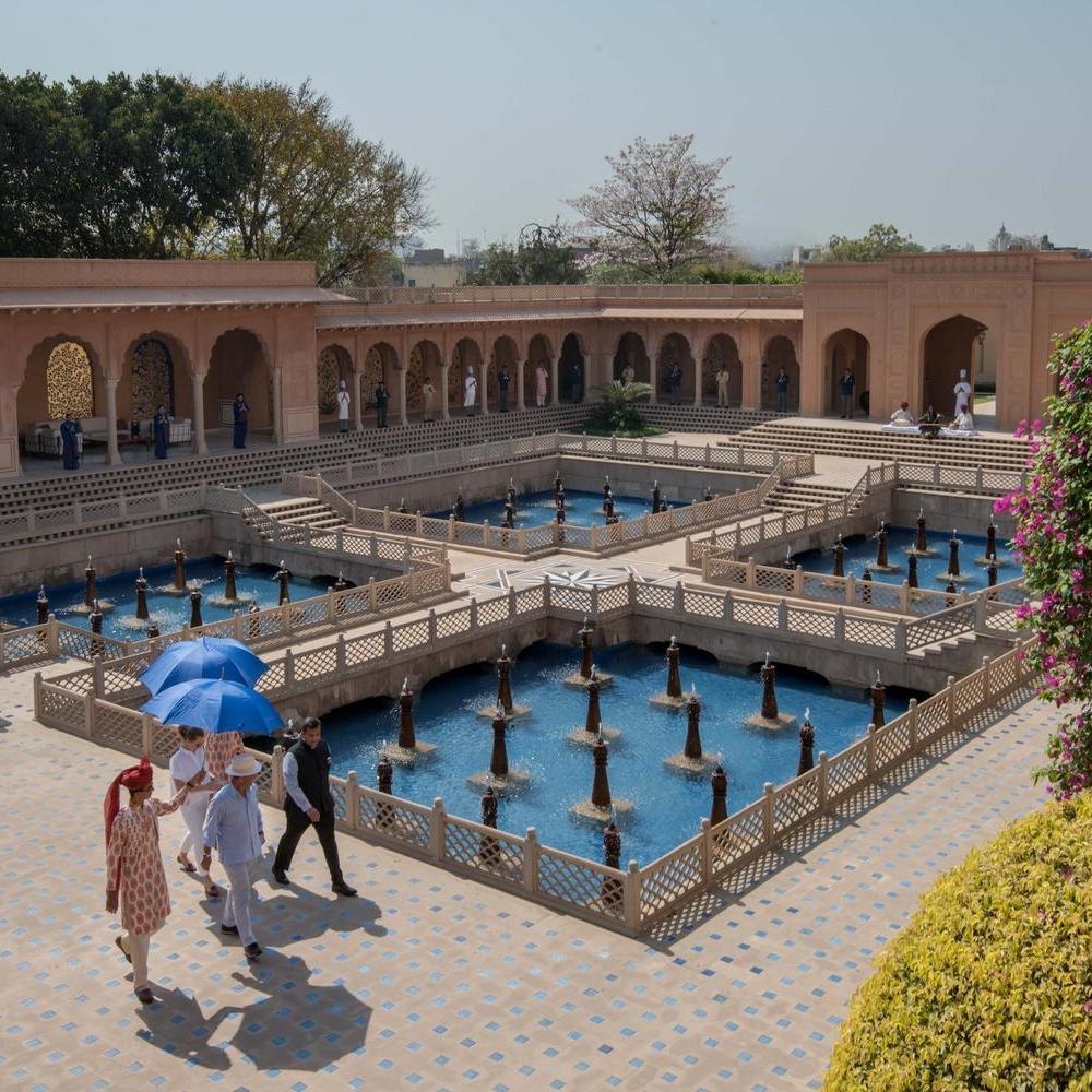 People walking past stepped reflection pools with fountains in a grand sandstone courtyard at The Oberoi Amarvilas.