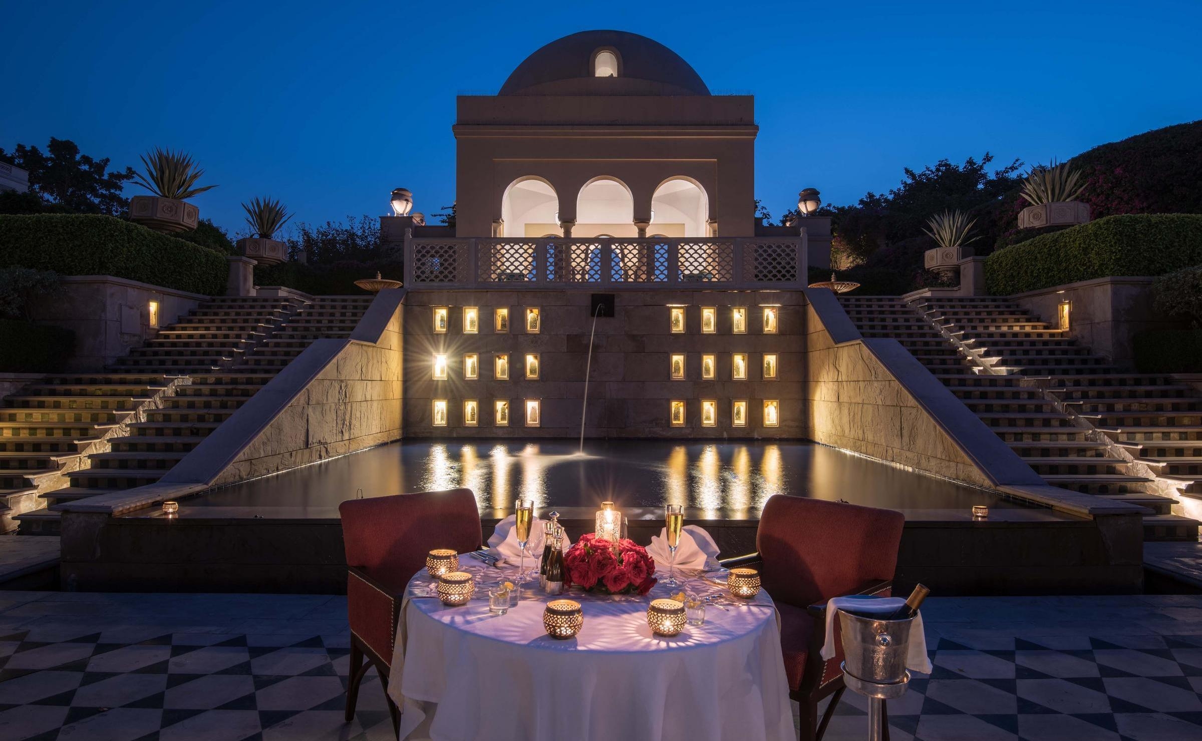 A candlelit dinner table for two by a fountain and illuminated stone building at night.