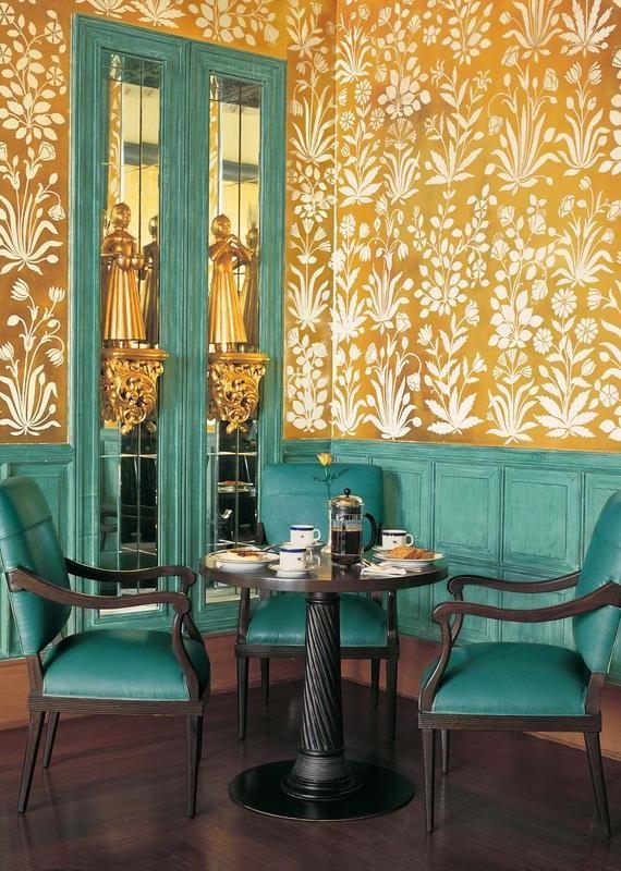 A small dining table with teal chairs against gold floral patterned walls and mirrored panels.