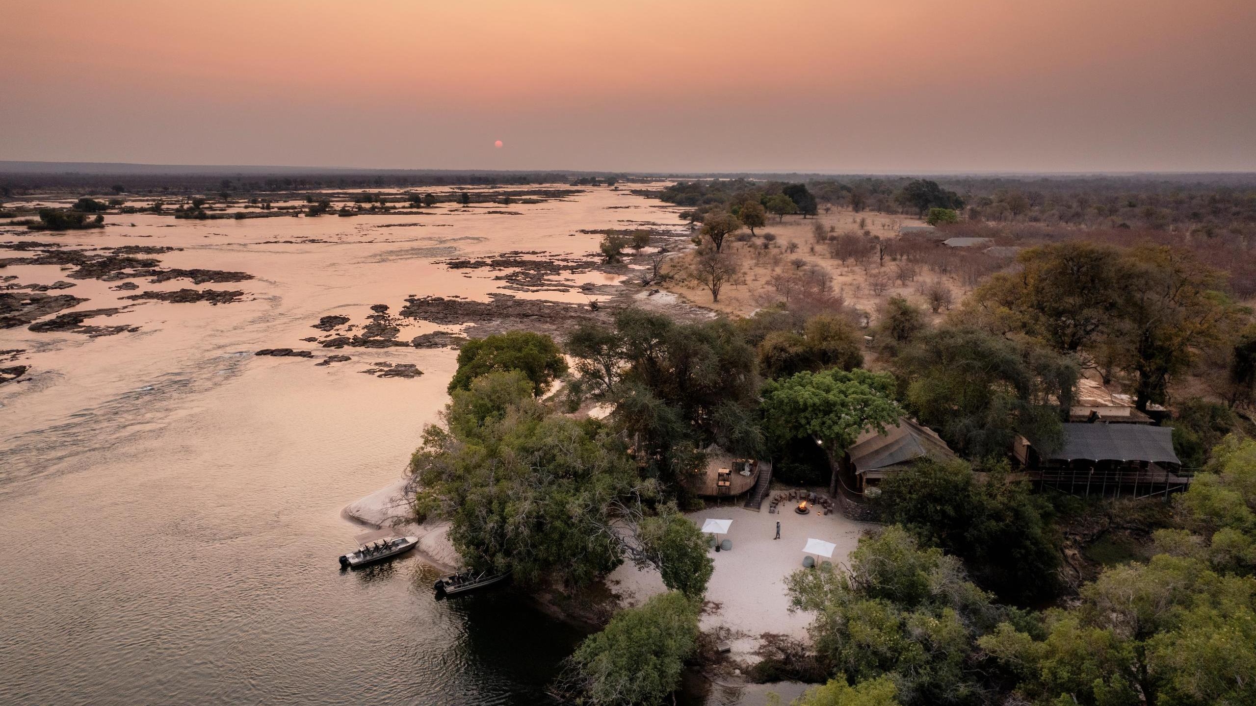Aerial view of a wide river at sunset with rocky rapids, a safari lodge among trees, and two small boats moored on the sand.