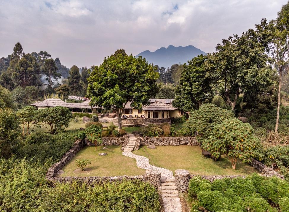 A stone pathway leading toward Mount Gahinga Lodge, surrounded by green trees and stone walls with mountains behind.