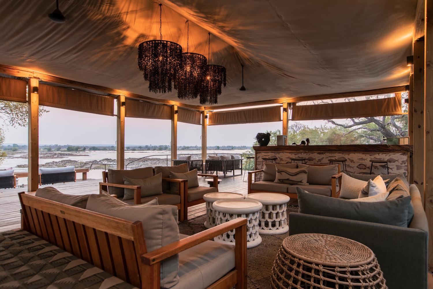 Open-air lounge under a canvas canopy featuring beaded chandeliers, plush wooden seating, a bar, and a view of a wide river.