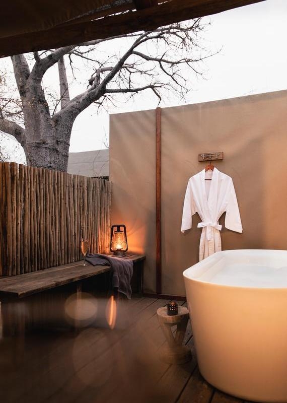 Outdoor wooden deck with a white bathtub, a hanging bathrobe, and a lit lantern and champagne glass on a rustic bench.