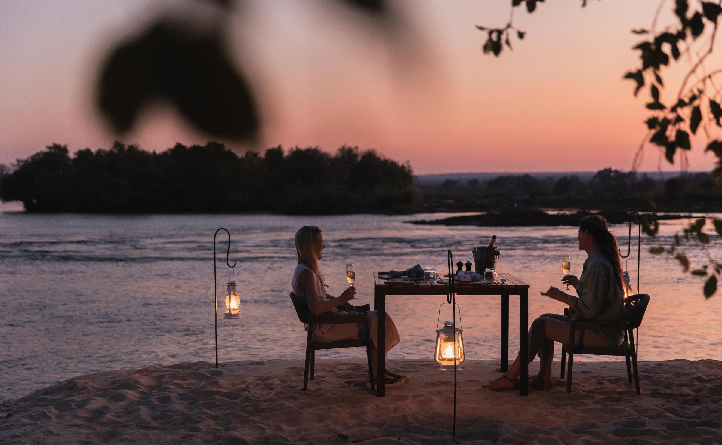 Two people enjoying a candlelit dinner at a table on a sandy riverbank at sunset, surrounded by glowing lanterns.