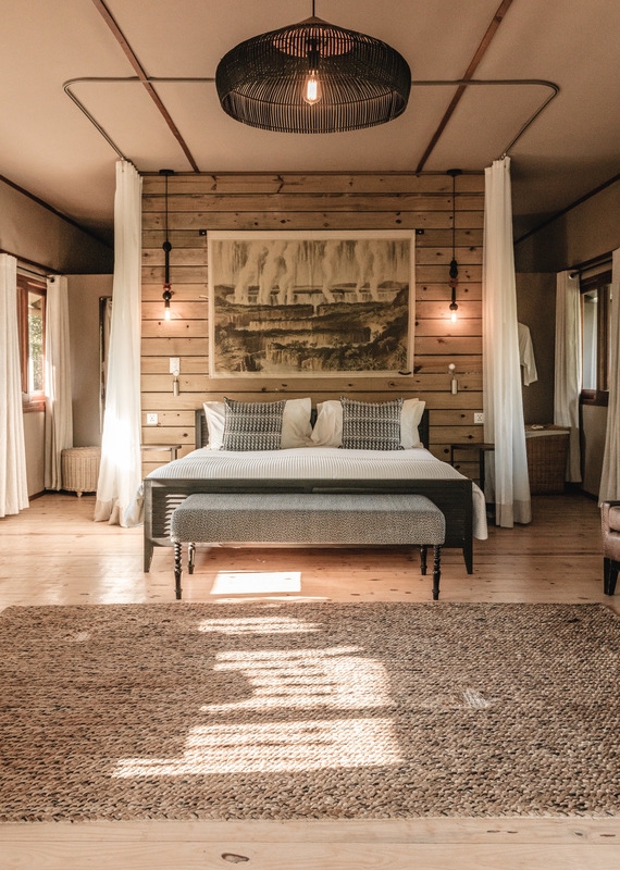 Cozy rustic bedroom featuring a canopy bed, a large nature tapestry, a black wicker chandelier, and a woven floor rug.