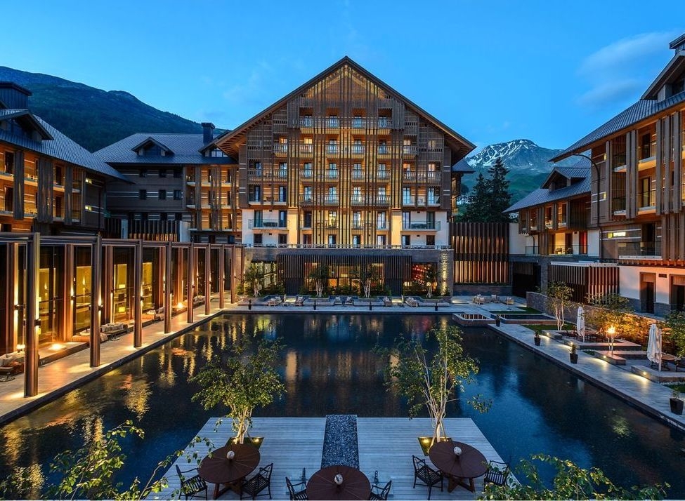 The Chedi Andermatt hotel courtyard and reflection pool at dusk with snow-capped mountains in the background.