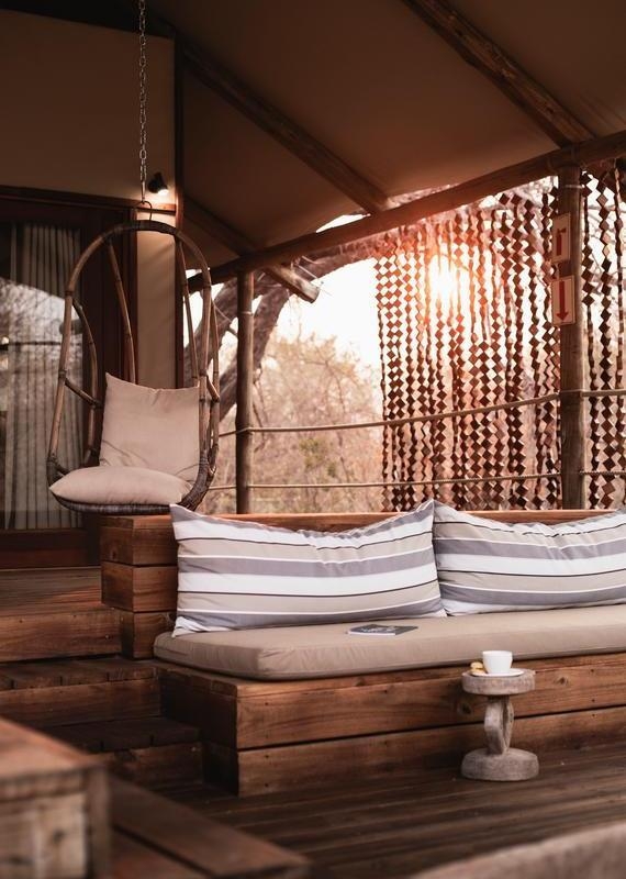 Shaded wooden deck at Toka Leya Camp with a hanging chair, daybed with striped pillows, and a small stool holding a cup.