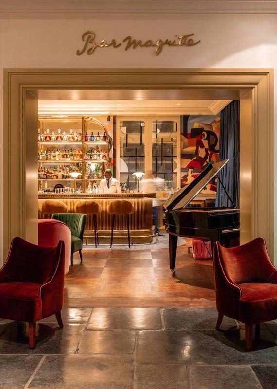 Entrance to Bar Magritte at hotel amigo in belgium, showing a piano, red chairs, and a bartender at a gold bar.