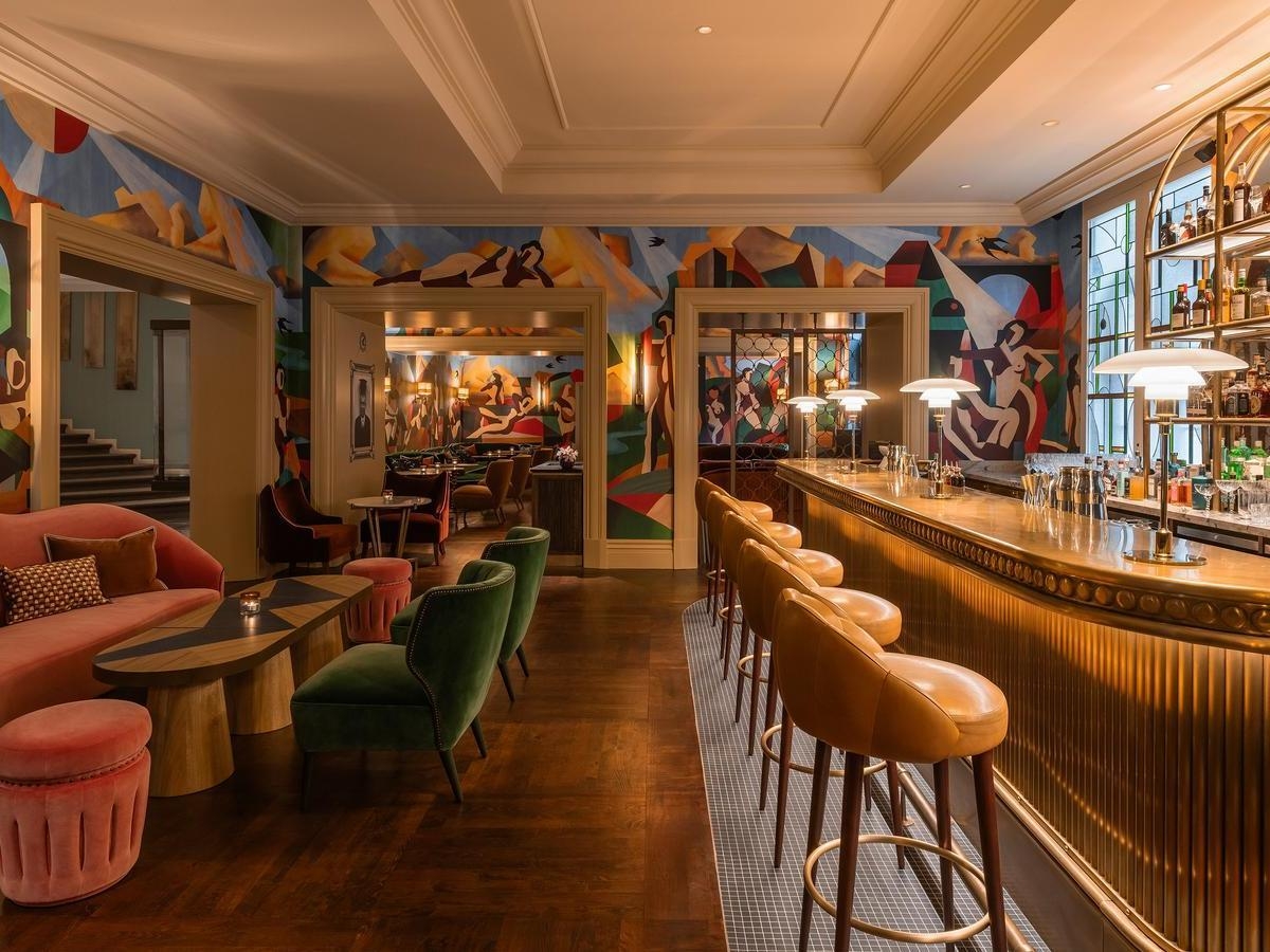 Elegant interior of Bar Magritte at hotel amigo in belgium featuring a gold bar, colorful murals, and velvet seating.