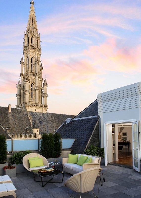 Rooftop terrace at hotel amigo in belgium with lounge seating and a view of a gothic spire at sunset.