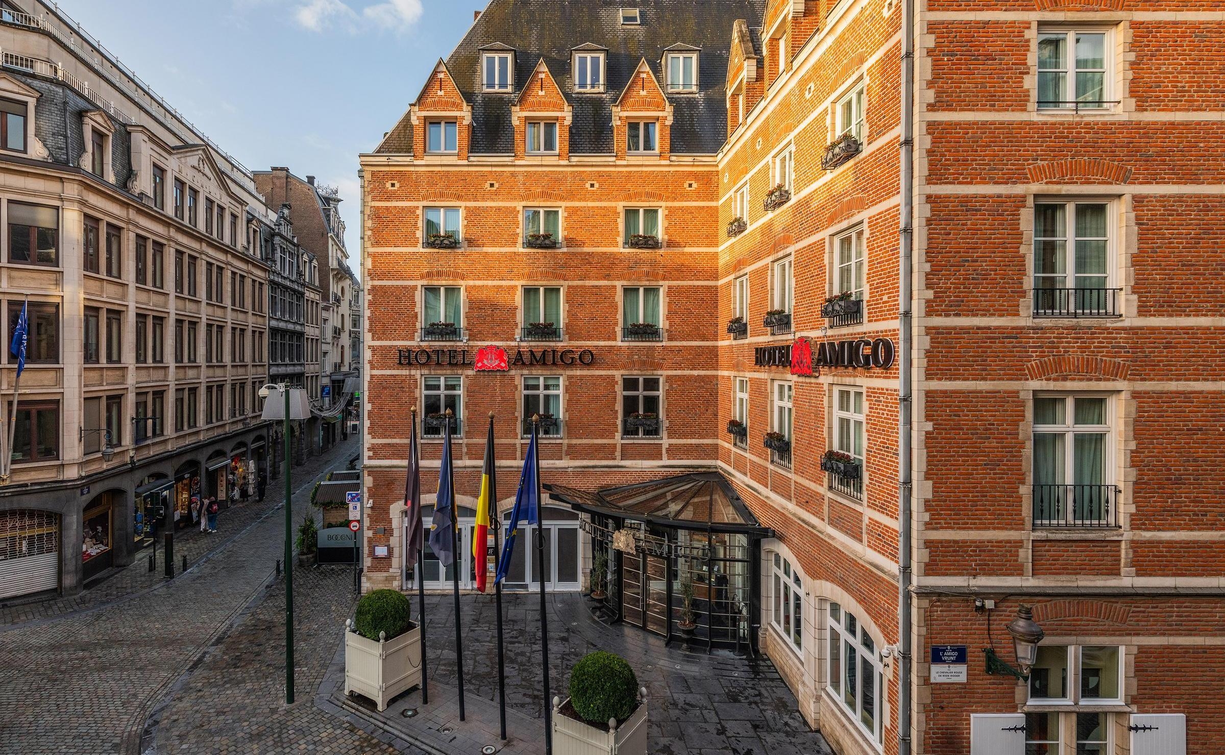 Red brick exterior of hotel amigo in belgium on a cobblestone street with flags and the hotel sign above the entrance.