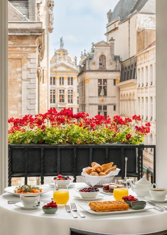 Breakfast table at hotel amigo in belgium looking through a window with red flowers toward historic city buildings.
