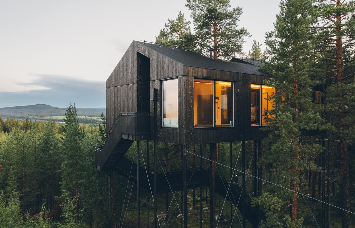 A large black wooden structure on high stilts surrounded by trees under a grey sky.
