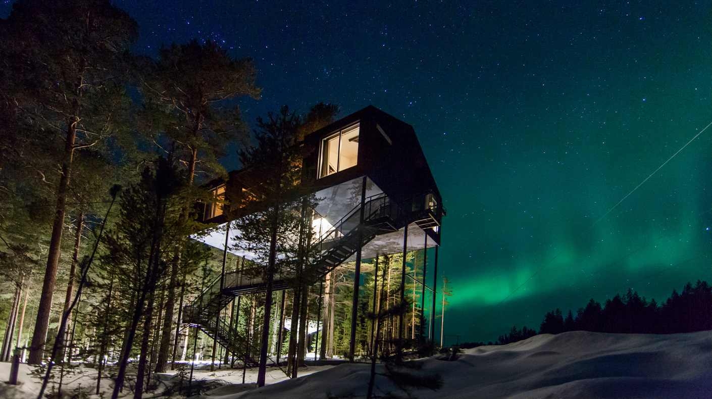 A modern dark cabin on high stilts illuminated against a starry sky with green northern lights.