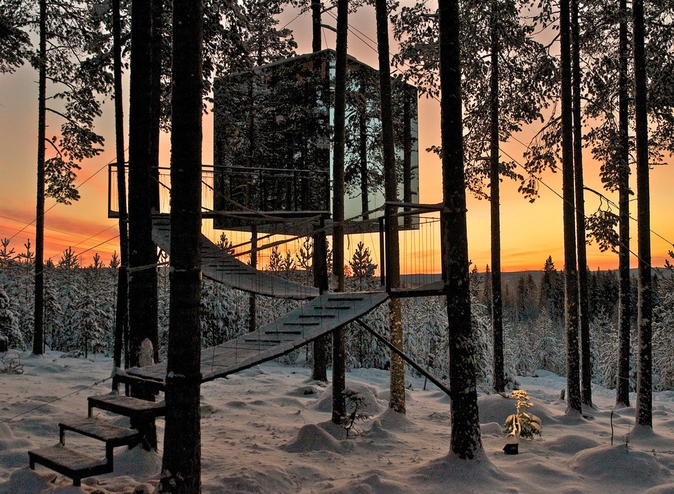 A square mirrored structure suspended in trees reflecting a snowy forest under a sunset sky.