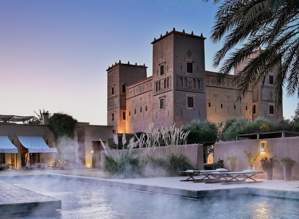 A steaming swimming pool at dusk with a large, lit-up Moroccan kasbah and palm trees in the background.