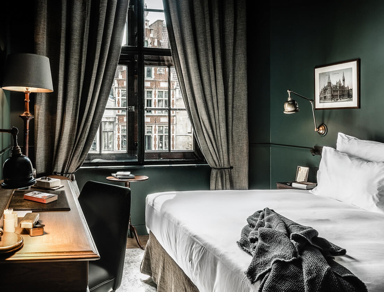 A bedroom with dark green walls, a white bed, a desk, and a window with curtains looking out at historic brick buildings.