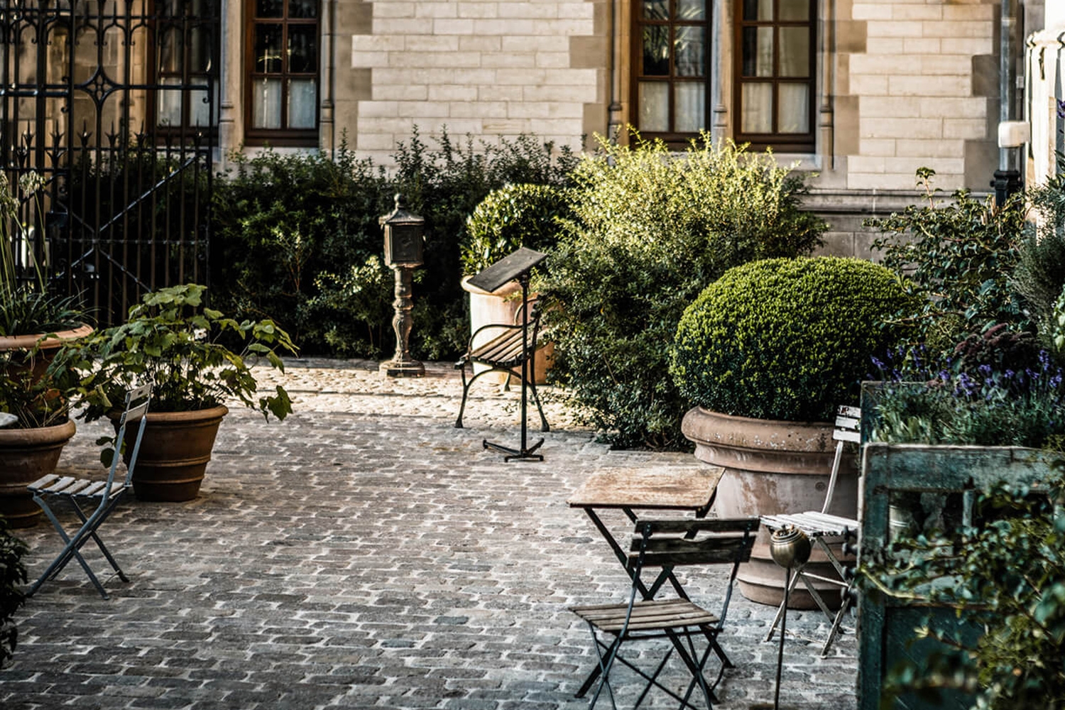 A historic cobblestone courtyard featuring potted plants, folding chairs, a small table, and a wooden bench.