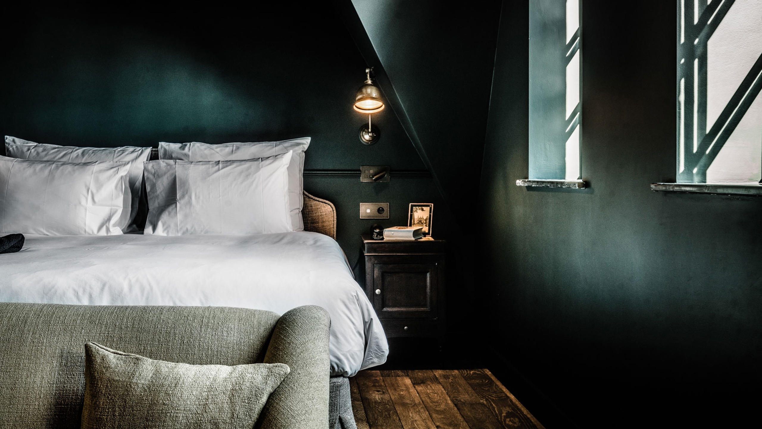 A hotel bedroom with dark green walls, a white bed, a small bedside table, a brass reading light, and a sloped ceiling.