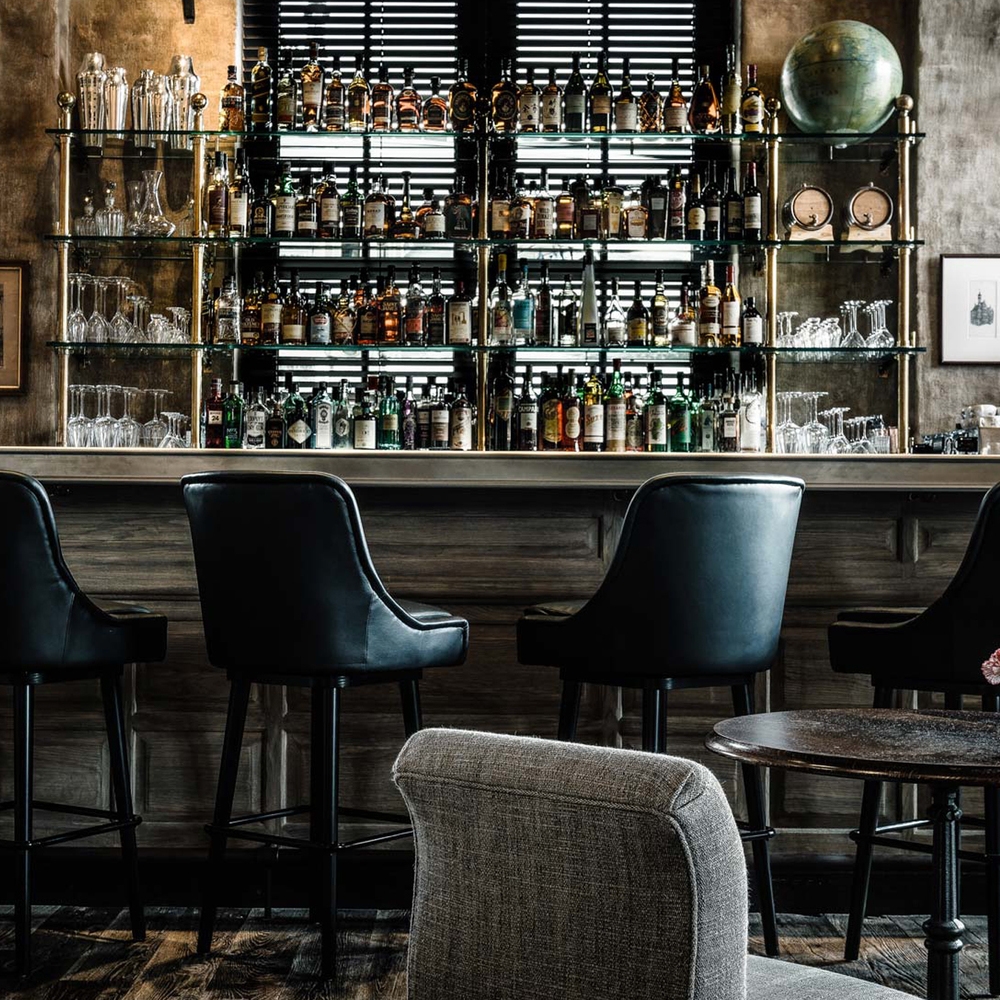 A dark cocktail bar featuring backlit glass shelves stocked with liquor bottles in front of a window, and leather bar stools.