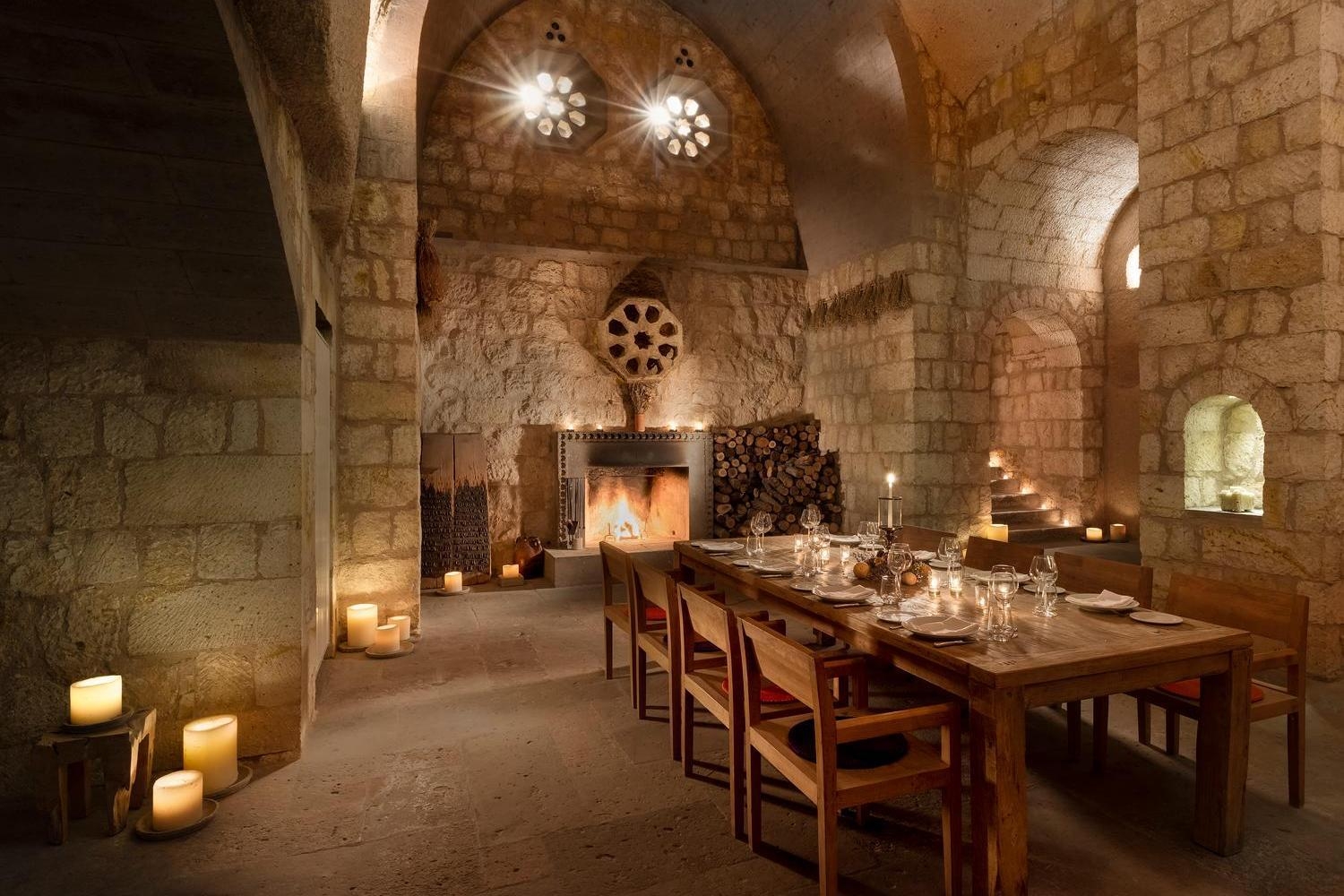 A long wooden dining table set for a meal inside a grand stone cave hall with candles.