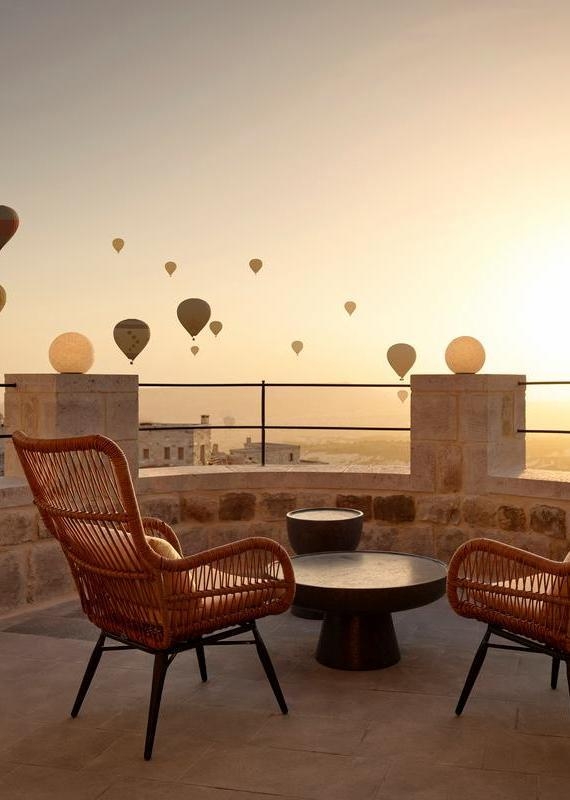 Wicker chairs on a stone terrace overlooking a valley filled with hot air balloons at dawn.